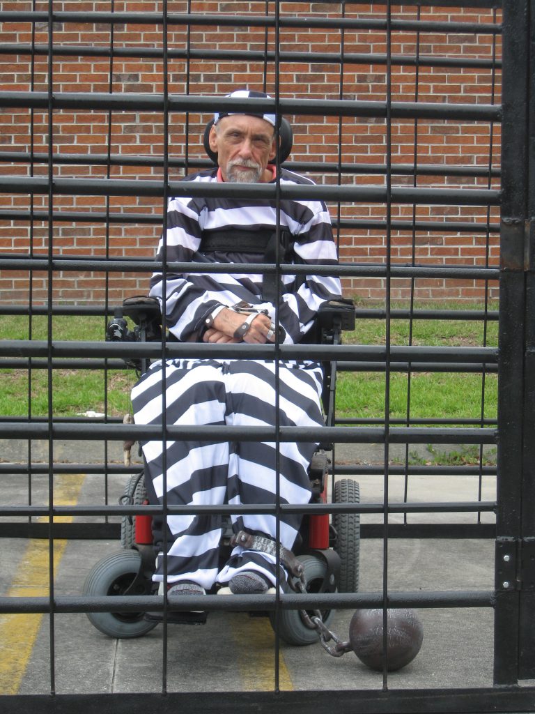 Picture of Bob Habas behind prison bars and dressed like a prisoner
